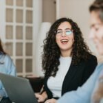 Three executives work on laptops at a table, smiling and talking together.